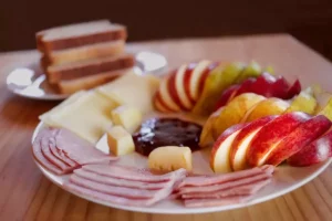 Desayuno tipo americano con un plato de frutas cortadas, jamón, queso, pan y mantequilla